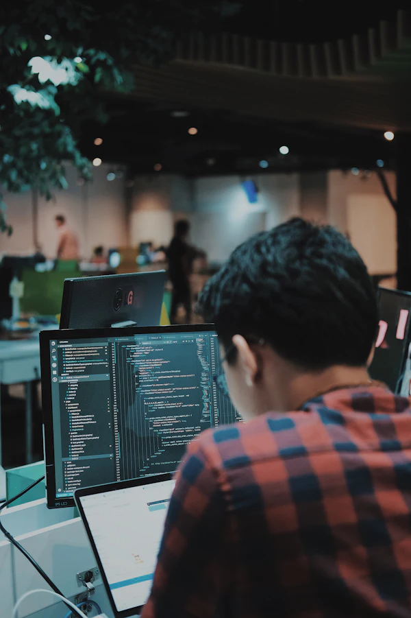 Developer sedang menulis kode di depan dua monitor di ruang kerja